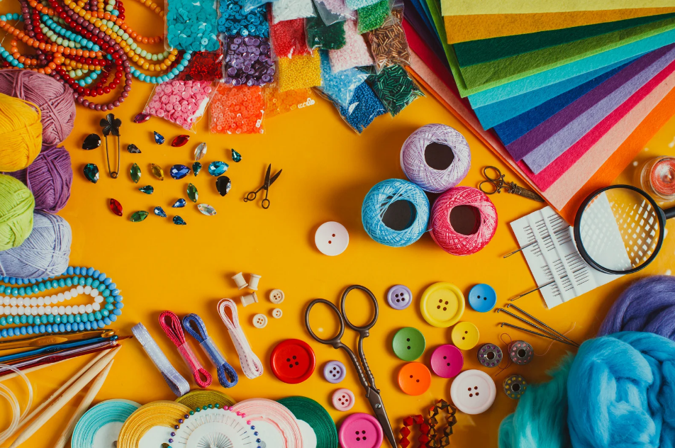 Crafting supplies arranged on a bright orange background including colorful buttons, yarn balls, beaded necklaces, scissors, felt sheets in rainbow colors, small bags of beads, and crafting tools. The composition features vibrant turquoise, pink, and yellow hues with organized clusters of similar items.