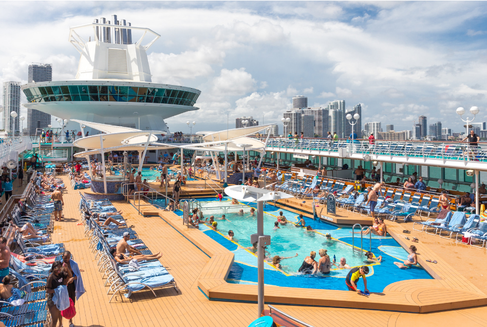 Cruise ship pool deck with swimming pools, lounge chairs, and Miami skyline in background